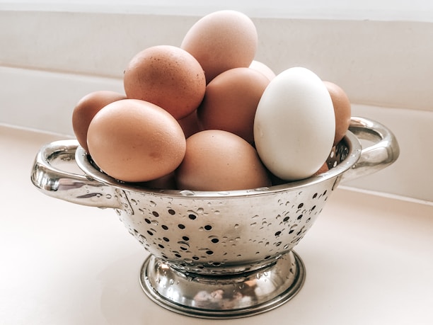A collection of eggs sits in a metal colander on a light-colored surface. The colander has two handles and features small holes scattered across its surface. The eggs vary in color, with most being brown and one distinctly white.
