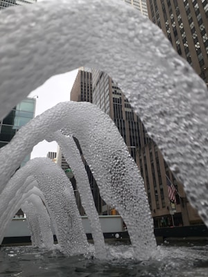 Arched streams of water are prominently displayed in front of tall city buildings, creating a dynamic urban water feature. The perspective shows water arcs in the foreground, with the skyscrapers forming the background. An American flag is visible, adding a touch of identity to the setting.