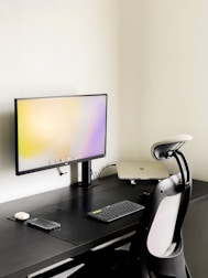 A modern minimalist workspace featuring a sleek black desk against a light-colored wall. A large computer monitor displays a light gradient screen and is placed centrally on the desk. Adjacent to the monitor is a closed laptop, slightly raised on a stand. A wireless keyboard and mouse are positioned near the front of the desk, alongside a smartphone. An ergonomic office chair with a mesh backrest is drawn up to the desk.