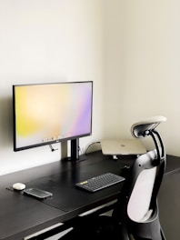 A modern minimalist workspace featuring a sleek black desk against a light-colored wall. A large computer monitor displays a light gradient screen and is placed centrally on the desk. Adjacent to the monitor is a closed laptop, slightly raised on a stand. A wireless keyboard and mouse are positioned near the front of the desk, alongside a smartphone. An ergonomic office chair with a mesh backrest is drawn up to the desk.