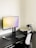A modern minimalist workspace featuring a sleek black desk against a light-colored wall. A large computer monitor displays a light gradient screen and is placed centrally on the desk. Adjacent to the monitor is a closed laptop, slightly raised on a stand. A wireless keyboard and mouse are positioned near the front of the desk, alongside a smartphone. An ergonomic office chair with a mesh backrest is drawn up to the desk.
