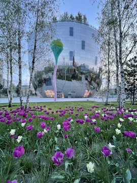 A futuristic, reflective glass building is surrounded by trees with a foreground of vibrant purple and white tulips. The structure is spherical with a mirrored surface, reflecting its surroundings. A colorful, abstract sculpture stands near the building.
