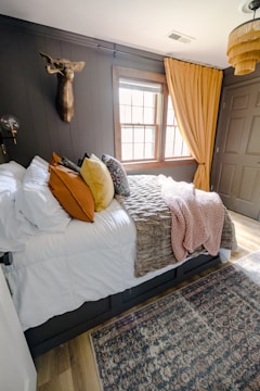 A cozy bedroom with a large bed covered in white sheets and various colored pillows including orange, yellow, and patterned designs. A textured throw blanket in brown and a pink knit throw are draped over the bed. The walls are dark, accented with a mounted decorative deer head and a modern light fixture. The window features wooden frames and yellow curtains, allowing natural light into the room. A woven basket-style ceiling lamp adds a touch of warmth, and a patterned area rug covers the wooden floor.