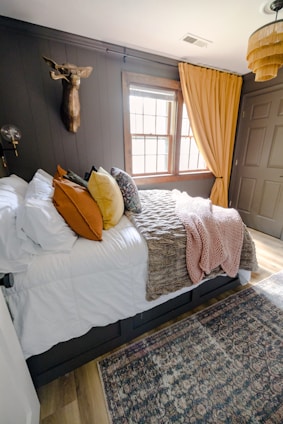 A cozy bedroom with a large bed covered in white sheets and various colored pillows including orange, yellow, and patterned designs. A textured throw blanket in brown and a pink knit throw are draped over the bed. The walls are dark, accented with a mounted decorative deer head and a modern light fixture. The window features wooden frames and yellow curtains, allowing natural light into the room. A woven basket-style ceiling lamp adds a touch of warmth, and a patterned area rug covers the wooden floor.