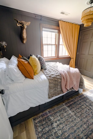 A cozy bedroom with a large bed covered in white sheets and various colored pillows including orange, yellow, and patterned designs. A textured throw blanket in brown and a pink knit throw are draped over the bed. The walls are dark, accented with a mounted decorative deer head and a modern light fixture. The window features wooden frames and yellow curtains, allowing natural light into the room. A woven basket-style ceiling lamp adds a touch of warmth, and a patterned area rug covers the wooden floor.