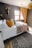 A cozy bedroom with a large bed covered in white sheets and various colored pillows including orange, yellow, and patterned designs. A textured throw blanket in brown and a pink knit throw are draped over the bed. The walls are dark, accented with a mounted decorative deer head and a modern light fixture. The window features wooden frames and yellow curtains, allowing natural light into the room. A woven basket-style ceiling lamp adds a touch of warmth, and a patterned area rug covers the wooden floor.
