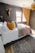 A cozy bedroom with a large bed covered in white sheets and various colored pillows including orange, yellow, and patterned designs. A textured throw blanket in brown and a pink knit throw are draped over the bed. The walls are dark, accented with a mounted decorative deer head and a modern light fixture. The window features wooden frames and yellow curtains, allowing natural light into the room. A woven basket-style ceiling lamp adds a touch of warmth, and a patterned area rug covers the wooden floor.