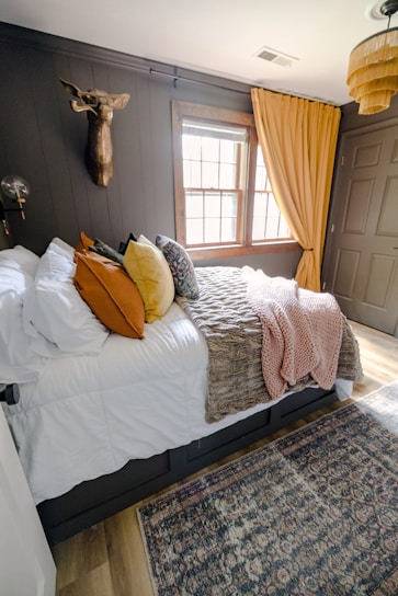 A cozy bedroom with a large bed covered in white sheets and various colored pillows including orange, yellow, and patterned designs. A textured throw blanket in brown and a pink knit throw are draped over the bed. The walls are dark, accented with a mounted decorative deer head and a modern light fixture. The window features wooden frames and yellow curtains, allowing natural light into the room. A woven basket-style ceiling lamp adds a touch of warmth, and a patterned area rug covers the wooden floor.