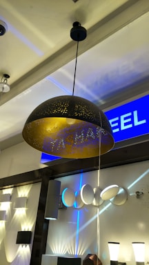 A modern lighting showroom featuring a variety of contemporary light fixtures. A large, dome-shaped pendant lamp with a black exterior and golden interior is the focal point. The background showcases an array of wall-mounted lights and lamps with diverse designs, contributing to a sophisticated and sleek atmosphere.