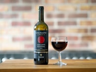 A bottle of red wine labeled 'Sagrato Chianti Riserva 2018' stands beside a filled wine glass, situated on a wooden surface with a blurred brick wall in the background.