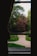 A landscaped garden view from a window featuring neatly trimmed shrubs and a gravel pathway leading through a lawn. Tall trees with varied shades of green and a tree with deep red leaves are visible in the background. The scene is framed by dark window edges, adding a sense of enclosure and perspective.