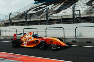 A bright orange and white Formula race car speeds along a racetrack, with an empty grandstand visible in the background. The car's aerodynamic design and sleek body are highlighted, and safety barriers line the track on both sides.