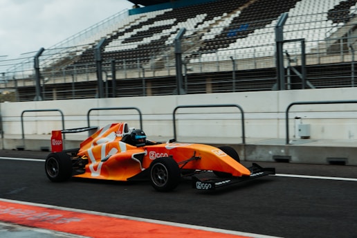 A bright orange and white Formula race car speeds along a racetrack, with an empty grandstand visible in the background. The car's aerodynamic design and sleek body are highlighted, and safety barriers line the track on both sides.