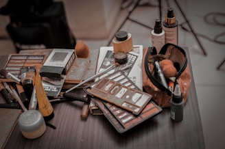 A collection of makeup items scattered on a tabletop, including various brushes, palettes, and bottles. A makeup bag filled with tools and products is prominently displayed, surrounded by eyeshadow and foundation containers.