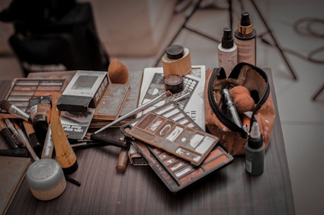 A collection of makeup items scattered on a tabletop, including various brushes, palettes, and bottles. A makeup bag filled with tools and products is prominently displayed, surrounded by eyeshadow and foundation containers.