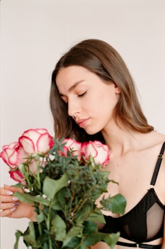 A young woman with long brown hair is holding a bouquet of roses. The roses are white with pink edges, and there is lush green foliage surrounding them. She is wearing a black top and has a serene expression with closed eyes.