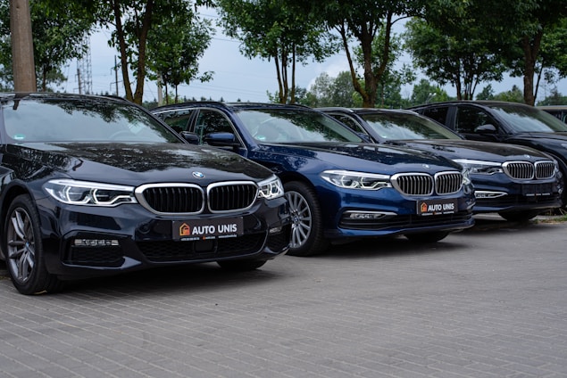 Several luxury sedans are parked in a row on a paved surface. The cars, which are dark in color, are positioned in a neatly organized manner under a canopy of green trees.
