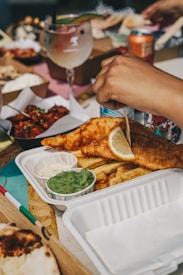 A hand is squeezing a lemon over a serving of fish and chips in a disposable white tray. Cups of mushy peas and tartar sauce accompany the meal. In the background, there are other food items and a large glass with a beverage garnished with fruit. The setting appears casual and outdoors with an assortment of colorful and diverse food options visible.