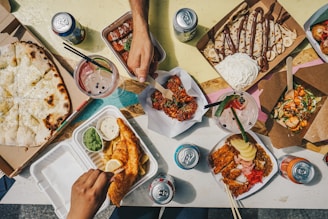 A flat lay of assorted foods on a table. The spread includes a large cheesy pizza, crispy fried chicken with a side of dipping sauces, a container of spicy sausages, a box of sweet crepes drizzled with chocolate and cream, a bowl of colorful salad with grilled meat, and several cans of soda. Two people are reaching in with chopsticks, ready to eat.