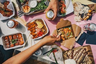 A table full of diverse and colorful food dishes, including fried chicken, salads, crepes topped with chocolate, and several canned drinks. Hands are reaching for food, indicating an informal dining setting.