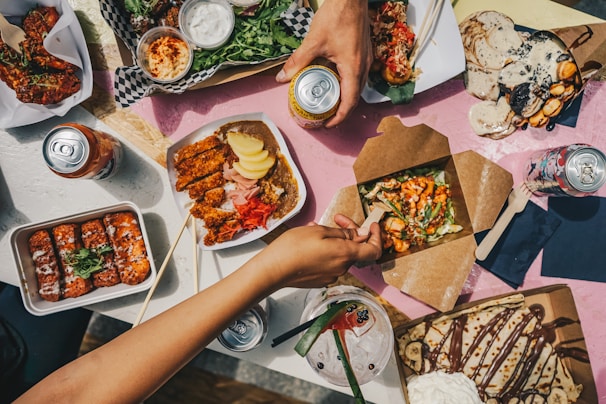 A table full of diverse and colorful food dishes, including fried chicken, salads, crepes topped with chocolate, and several canned drinks. Hands are reaching for food, indicating an informal dining setting.