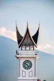 A clock tower with traditional Minangkabau architecture, featuring a distinctive curved roof with pointed tips. The clock face is centered on the white tower, and the background includes a hazy view of a mountain range against a partly cloudy sky.