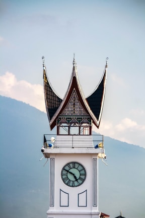 A clock tower with traditional Minangkabau architecture, featuring a distinctive curved roof with pointed tips. The clock face is centered on the white tower, and the background includes a hazy view of a mountain range against a partly cloudy sky.