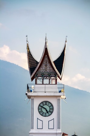 A clock tower with traditional Minangkabau architecture, featuring a distinctive curved roof with pointed tips. The clock face is centered on the white tower, and the background includes a hazy view of a mountain range against a partly cloudy sky.