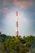 A tall communication tower with red and white sections stands prominently against a backdrop of clouds and a vast blue sky. The structure is surrounded by lush green trees, suggesting a remote or natural setting.