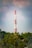A tall communication tower with red and white sections stands prominently against a backdrop of clouds and a vast blue sky. The structure is surrounded by lush green trees, suggesting a remote or natural setting.