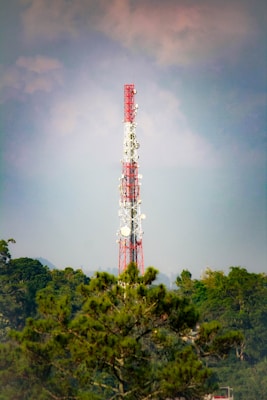 A tall communication tower with red and white sections stands prominently against a backdrop of clouds and a vast blue sky. The structure is surrounded by lush green trees, suggesting a remote or natural setting.