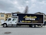 A large moving truck is parked on a residential street in front of a house with beige siding and white garage door. The truck is branded with 'Los Angeles Transfer And Storage,' featuring contact information and graphics of a city skyline. The sky is overcast, and the street is empty.