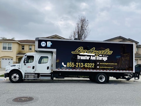 A large moving truck is parked on a residential street in front of a house with beige siding and white garage door. The truck is branded with 'Los Angeles Transfer And Storage,' featuring contact information and graphics of a city skyline. The sky is overcast, and the street is empty.