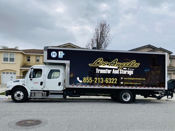 A large moving truck is parked on a residential street in front of a house with beige siding and white garage door. The truck is branded with 'Los Angeles Transfer And Storage,' featuring contact information and graphics of a city skyline. The sky is overcast, and the street is empty.