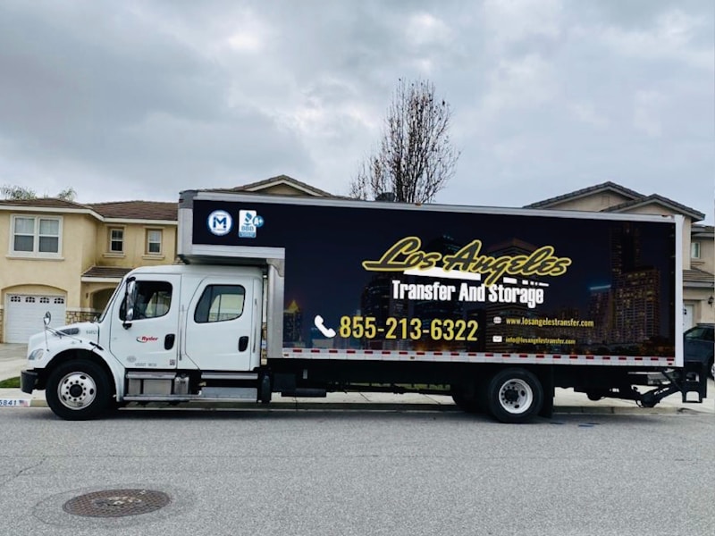 A large moving truck is parked on a residential street in front of a house with beige siding and white garage door. The truck is branded with 'Los Angeles Transfer And Storage,' featuring contact information and graphics of a city skyline. The sky is overcast, and the street is empty.