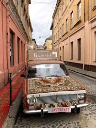 A vintage car is parked in a narrow cobblestone street, covered with an ornate Persian-style rug. The building facades on either side are painted in muted earth tones, and there is a sign on top of the car displaying 'Gazda Cafe' with text in Cyrillic script.
