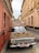 A vintage car is parked in a narrow cobblestone street, covered with an ornate Persian-style rug. The building facades on either side are painted in muted earth tones, and there is a sign on top of the car displaying 'Gazda Cafe' with text in Cyrillic script.
