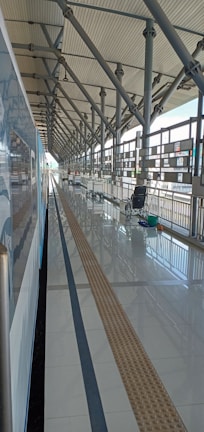 A modern train station platform with a train on the left side. The platform is clean, with reflective tiles, and features overhead structural beams and a metal railing. In the distance, there are a few empty luggage carts and a wheelchair near the railing.