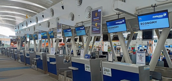 A series of check-in counters at an airport, specifically for economy class passengers. The counters have digital displays with flight information and signage for Malaysia Airlines. The architecture includes a modern design with high ceilings and plenty of natural light.
