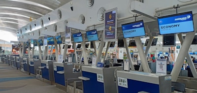 A series of check-in counters at an airport, specifically for economy class passengers. The counters have digital displays with flight information and signage for Malaysia Airlines. The architecture includes a modern design with high ceilings and plenty of natural light.
