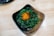 A square dish contains a serving of green seaweed salad topped with a small mound of bright orange fish roe. The bowl sits on a light wooden surface, creating a contrast with the dark dish and vibrant colors of the salad and roe.