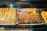 An assortment of colorful and intricately designed pastries displayed on metal trays. The pastries feature bright green, red, and creamy white decorations and are arranged neatly in rows. The scene is inside a bakery display case, lit with warm lighting.