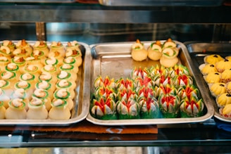 An assortment of colorful and intricately designed pastries displayed on metal trays. The pastries feature bright green, red, and creamy white decorations and are arranged neatly in rows. The scene is inside a bakery display case, lit with warm lighting.