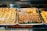 An assortment of colorful and intricately designed pastries displayed on metal trays. The pastries feature bright green, red, and creamy white decorations and are arranged neatly in rows. The scene is inside a bakery display case, lit with warm lighting.