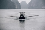 A solitary fishing boat equipped with long, outstretched arms for netting activities moves across a calm body of water. The scene is set against a backdrop of misty, towering cliffs, lending a serene and somewhat mysterious atmosphere to the image.