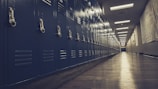 A long hallway features rows of blue metal lockers lining the walls on either side. The floor is made of polished wood, and the ceiling is fitted with fluorescent lighting, casting a subtle glow over the scene. The perspective stretches down the length of the corridor, creating a sense of depth and leading toward a softly lit doorway.