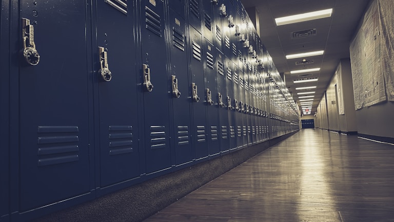 A long hallway features rows of blue metal lockers lining the walls on either side. The floor is made of polished wood, and the ceiling is fitted with fluorescent lighting, casting a subtle glow over the scene. The perspective stretches down the length of the corridor, creating a sense of depth and leading toward a softly lit doorway.