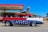 A vintage car designed with a patriotic theme featuring white stars on a blue background along the side, combined with red and white colors. A large cowboy statue is perched on the roof of the car. The car is parked in front of a vintage-style building with signs indicating Route 66 and souvenirs. There are a few people standing nearby, and an ice cream shop is visible in the background under a clear blue sky.