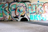 Colorful graffiti art covering a wall in an urban setting. The artwork features abstract shapes, swirling lines, and bold letters with a cartoonish face in the center. The vibrant colors include shades of blue, pink, orange, and green. The ground in front of the wall is a mixture of concrete and metal grating.