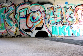 Colorful graffiti art covering a wall in an urban setting. The artwork features abstract shapes, swirling lines, and bold letters with a cartoonish face in the center. The vibrant colors include shades of blue, pink, orange, and green. The ground in front of the wall is a mixture of concrete and metal grating.